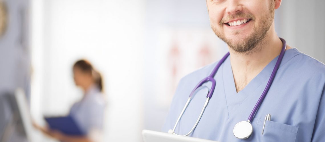 a mid adult male doctor stands with his touchscreen and smiles to camera . He is  dressed in blue medical scrubs , with a stethoscope around his neck , holding a digital tablet .In the background there is a defocussed nurse working at a computer.